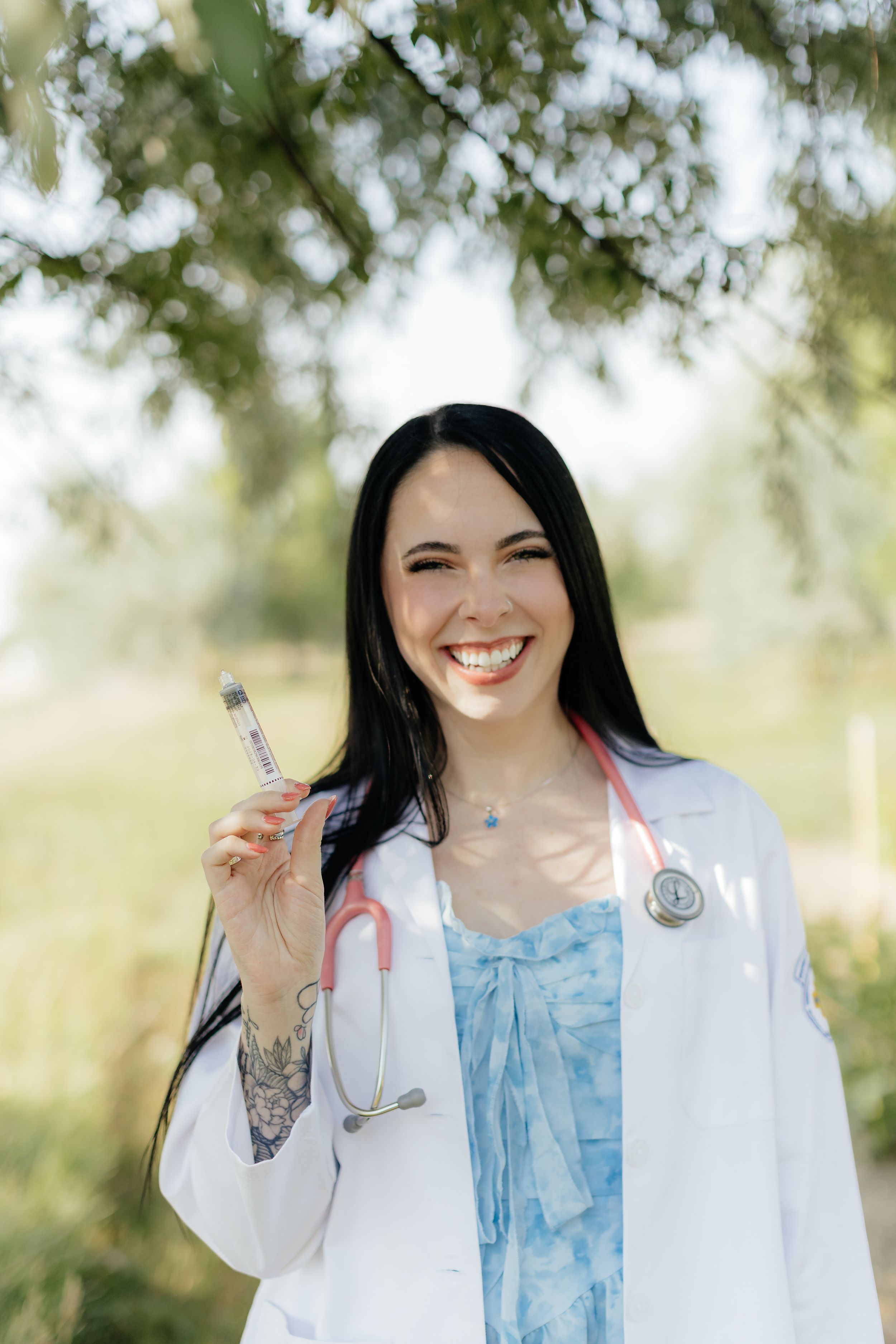 Smiling female doctor in white coat with pink stethoscope holding syringe outdoors under green trees.