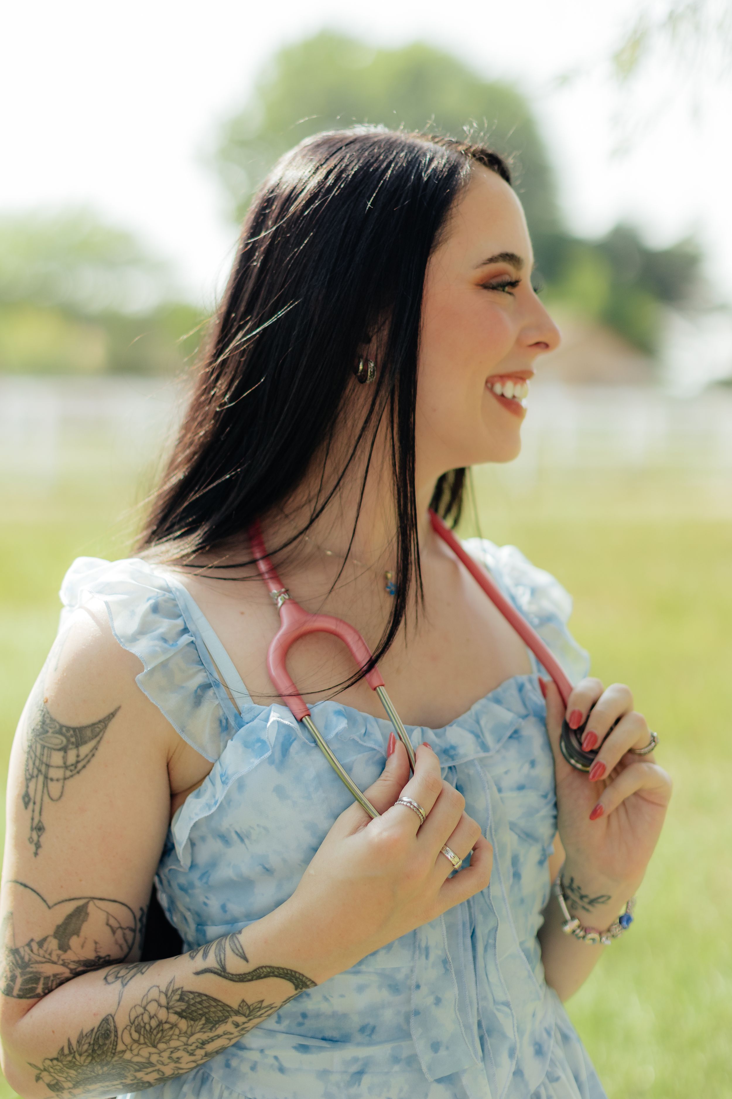Smiling woman with black hair and tattoos wearing a blue floral dress and pink stethoscope outdoors.