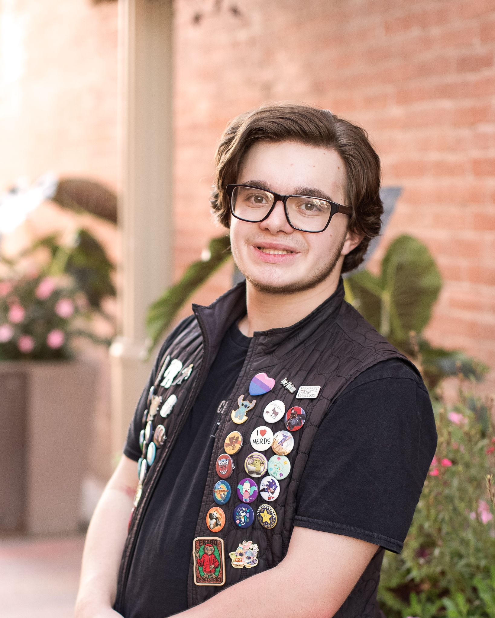 Student wearing glasses and a black vest adorned with colorful pins and patches stands near brick wall and plants.