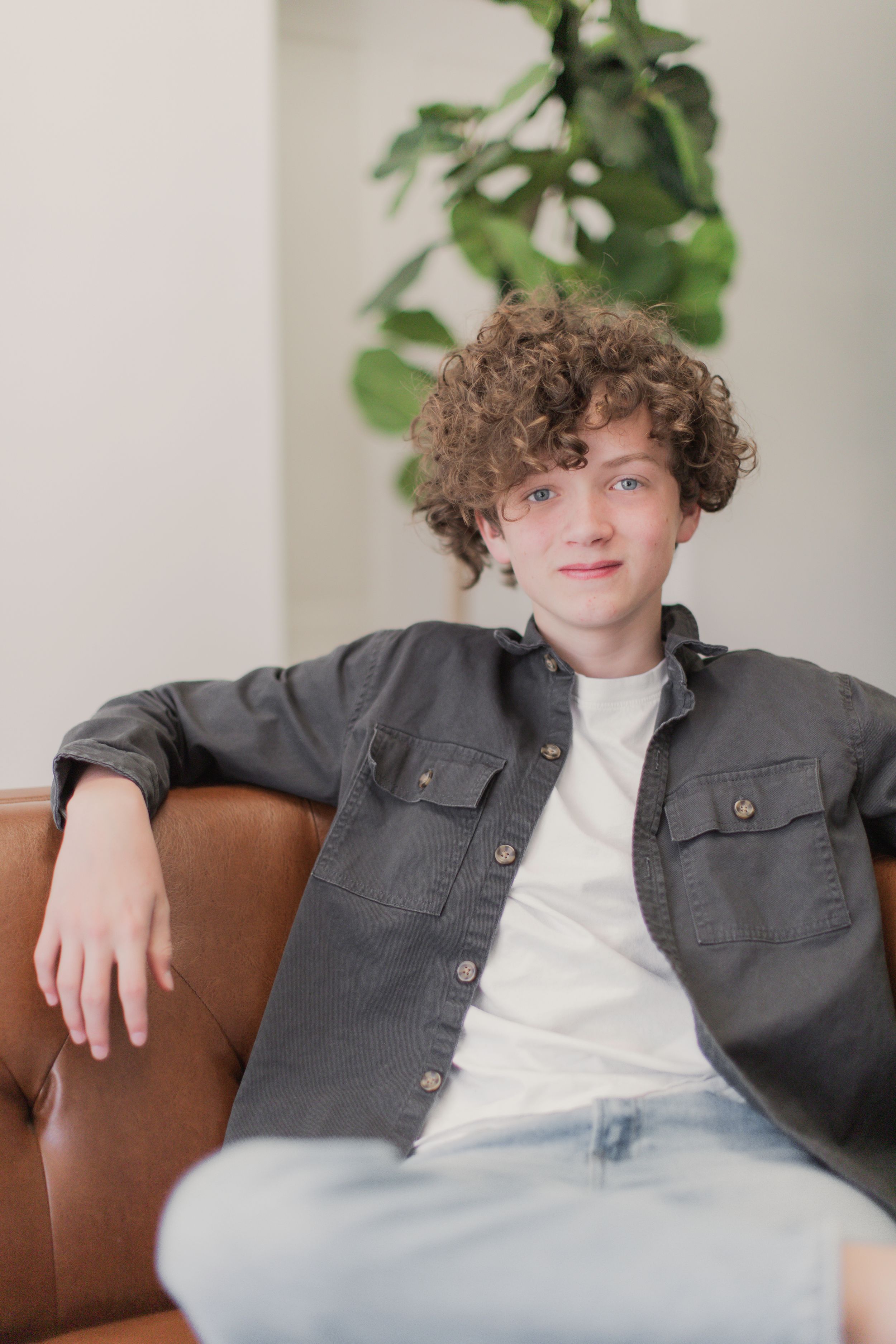 A young person with curly brown hair relaxes on a brown leather couch wearing a gray button-up shirt over white t-shirt.
