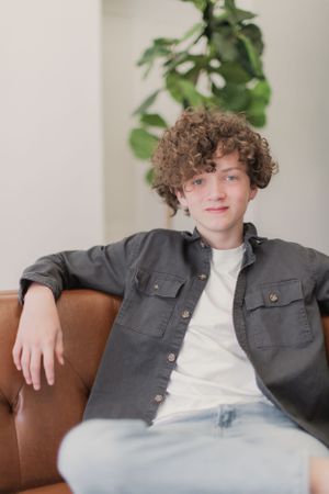 A young person with curly brown hair relaxes on a brown leather couch wearing a gray button-up shirt over white t-shirt.
