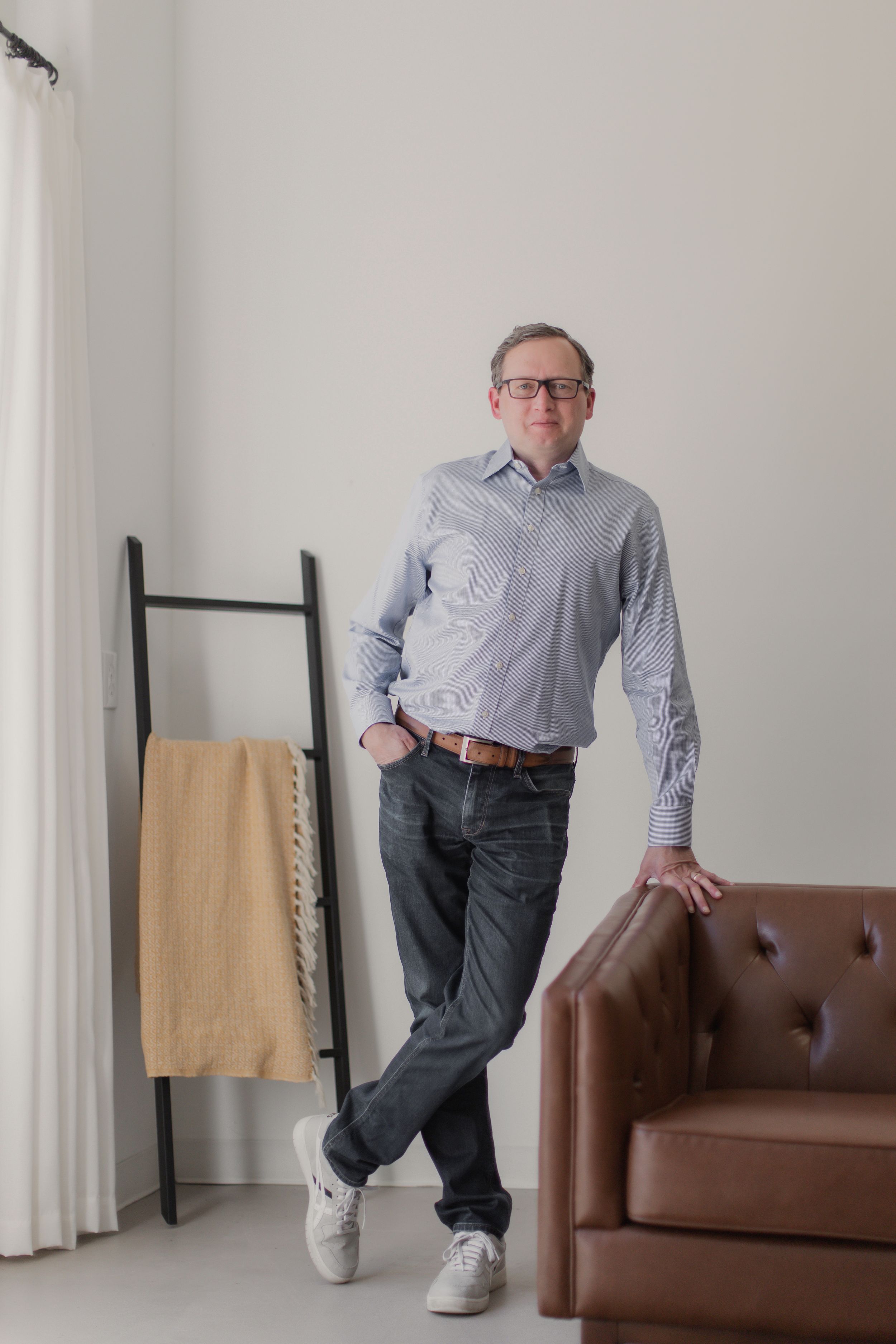 Mature adult wearing light blue dress shirt, jeans and white sneakers casually leaning against brown leather armchair.
