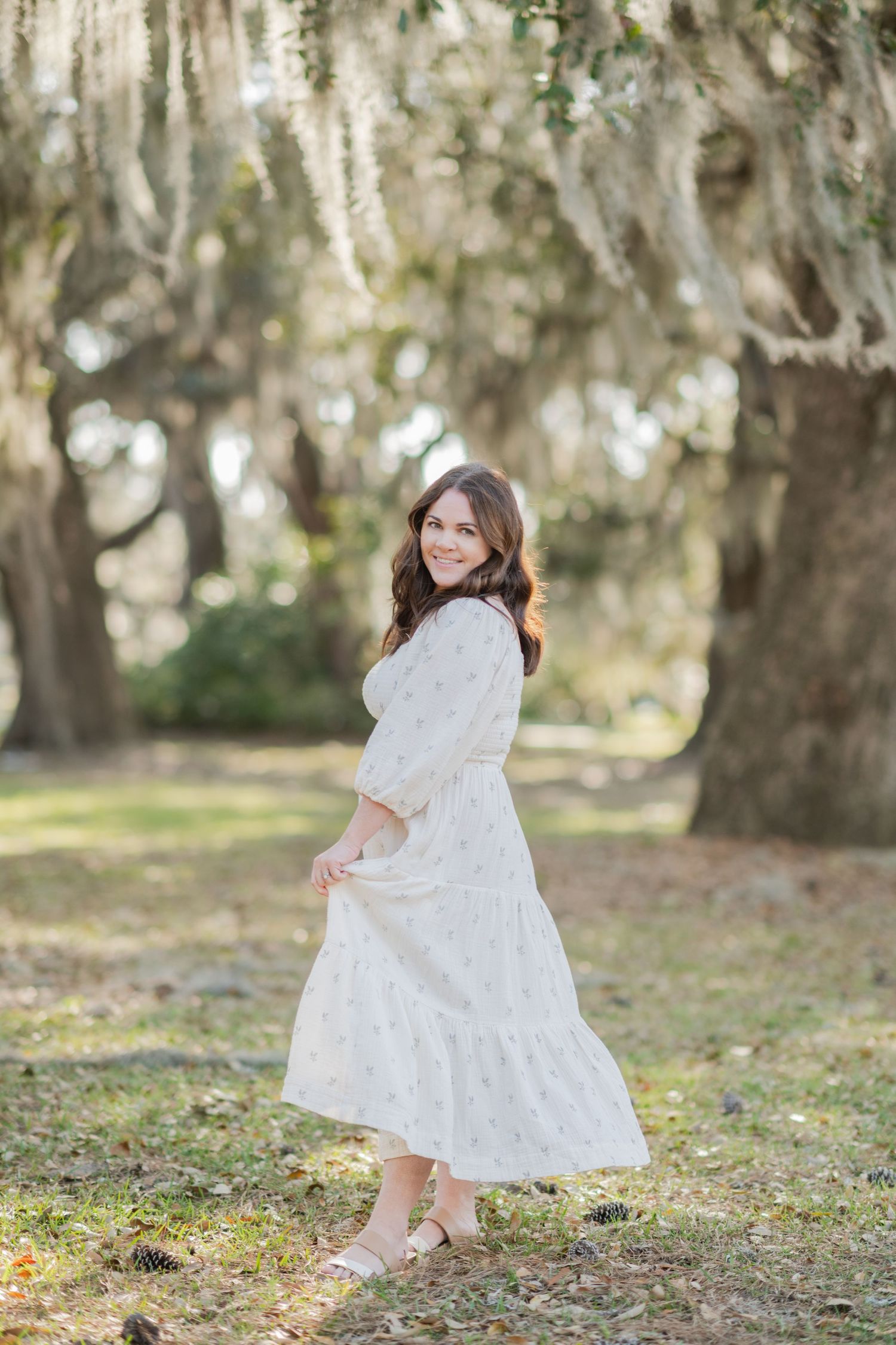 A woman in a white floral midi dress twirls under moss-draped oak trees in a park.