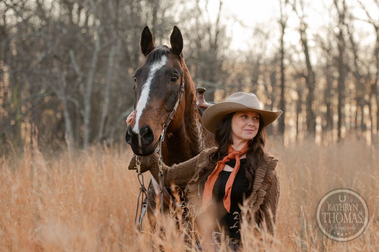 A horse and rider stand in tall autumn grass with bare trees behind them.