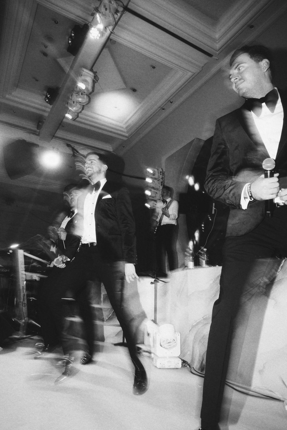 Motion-blurred black and white photo of performers in tuxedos at formal event.