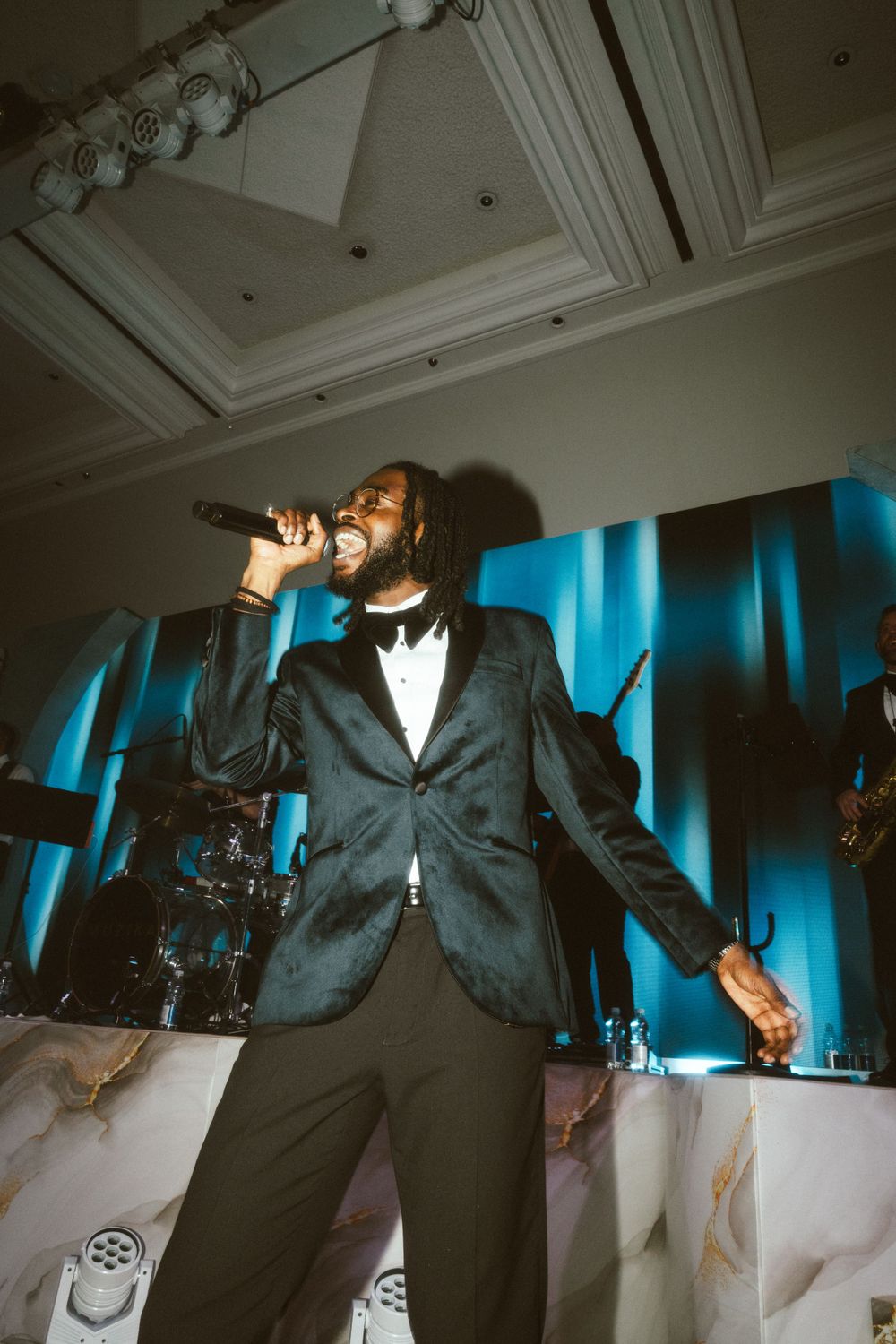 A performer in a black suit and bow tie sings passionately on stage with blue lighting.