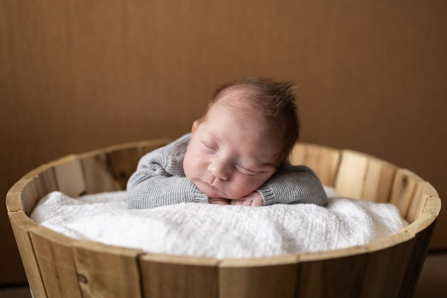 Sleeping newborn in grey outfit resting in wooden basket with white cushioning.