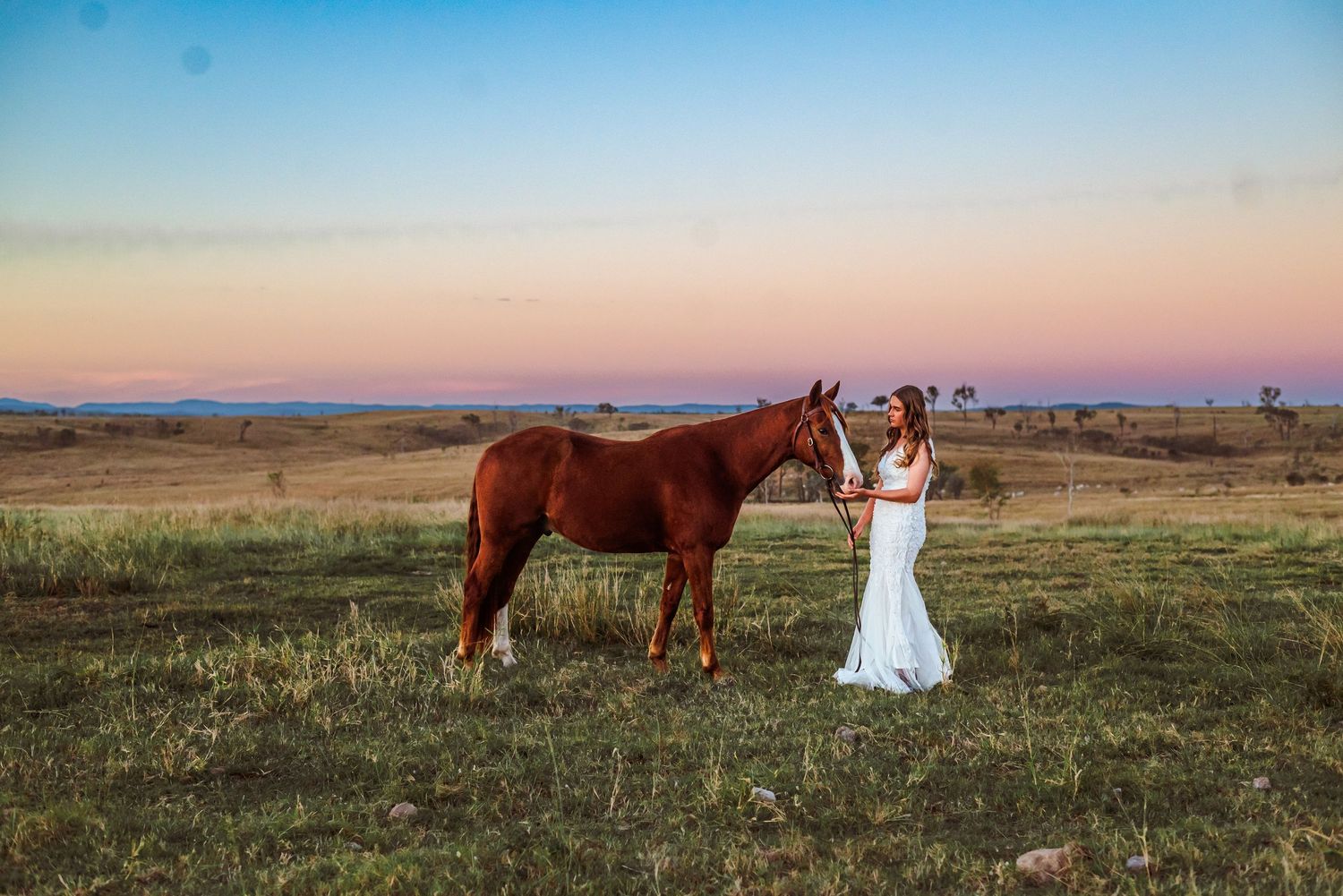 A brown horse and person in a white dress stand together in a field at sunset.