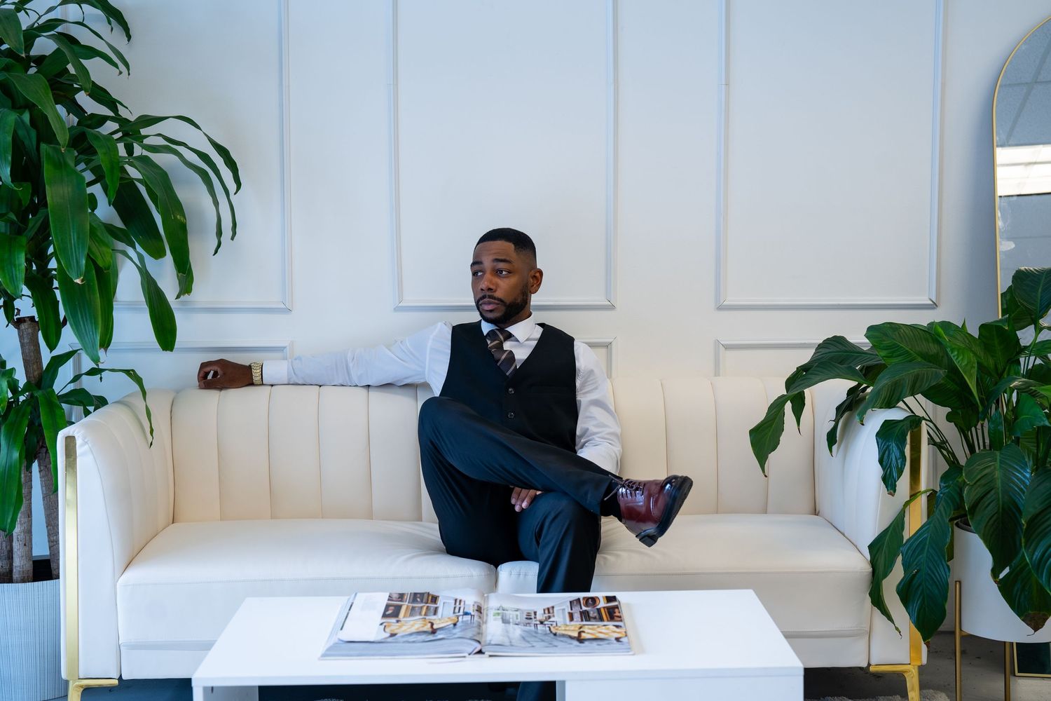 Someone sits on a white sofa wearing business attire with a magazine on the coffee table and plants in the background.