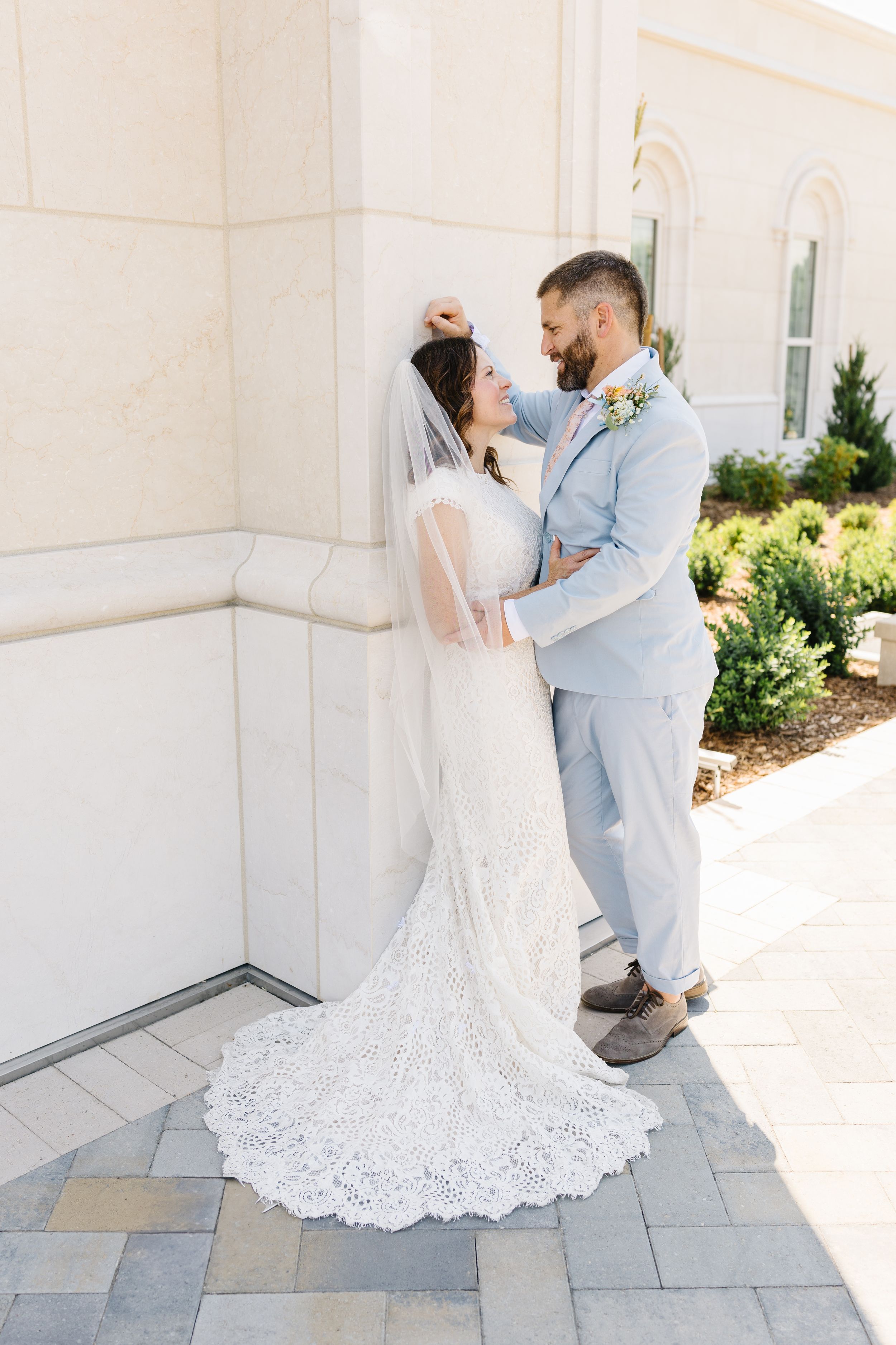 Couple in wedding attire embracing against white building exterior.