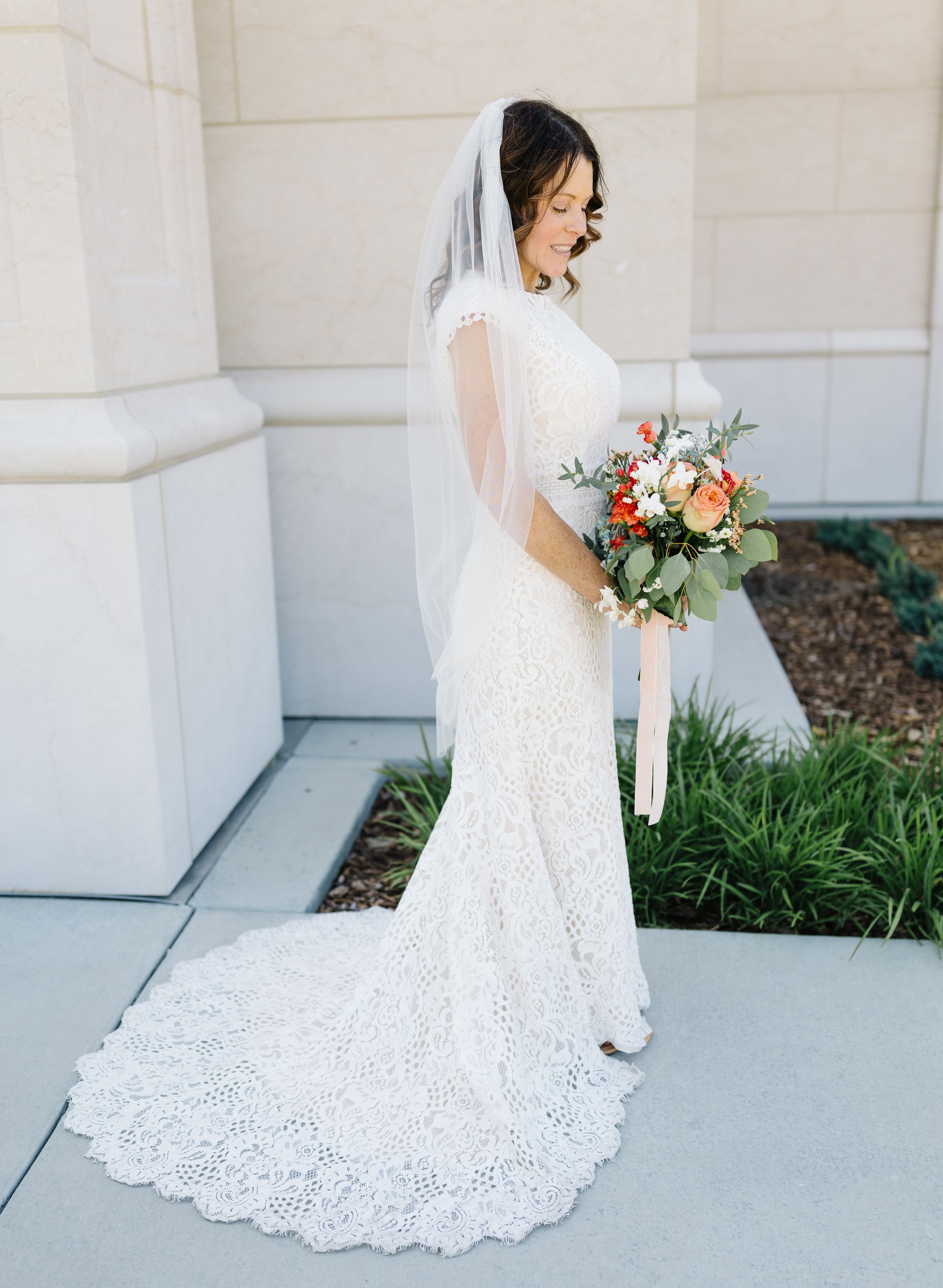 Bride in lace gown with long train holds peach and white bouquet.
