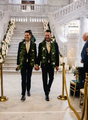 Wedding couple wearing matching dark suits and floral garlands walk down elegant marble staircase at venue.