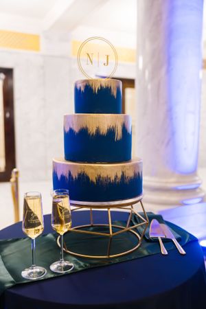 Three-tiered navy blue and gold wedding cake with monogram topper displayed with champagne glasses.