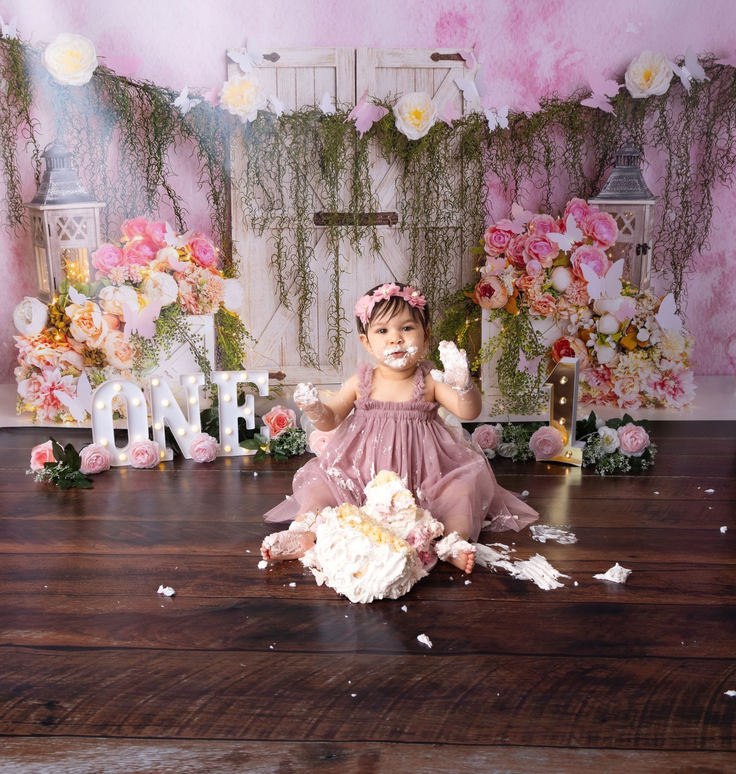 First birthday cake smash setup with pink florals and rustic white furniture against wooden floor.