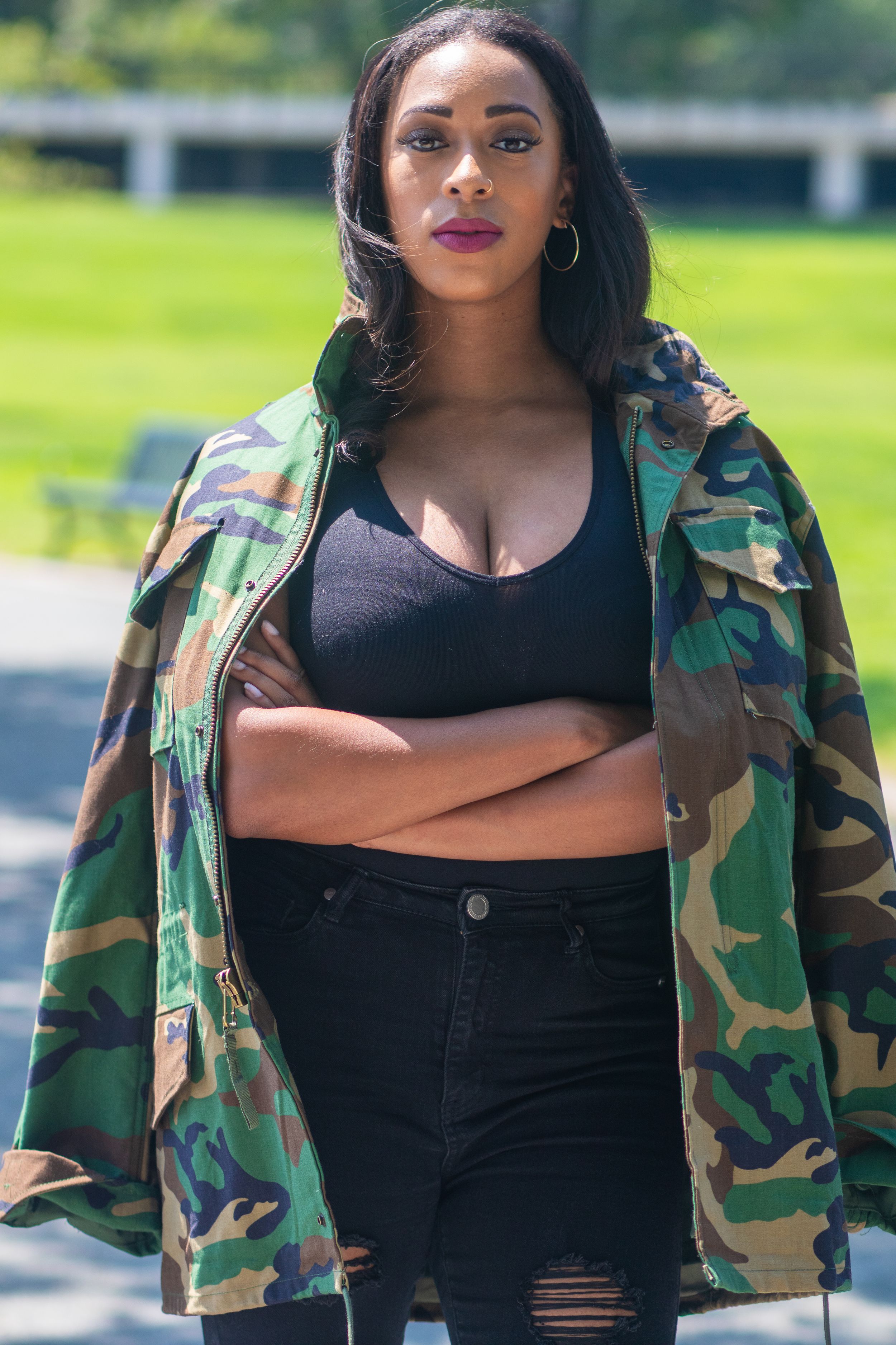 A stylish streetwear look with camouflage jacket, black crop top and distressed jeans against a green outdoor background.