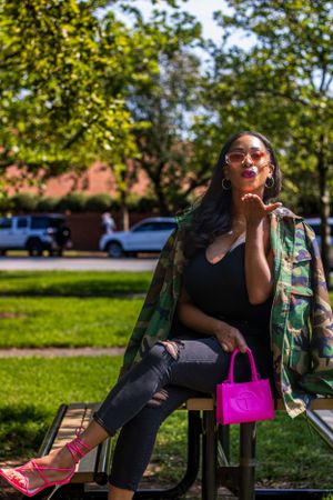 Fashion shot showing camo jacket with bright pink handbag and red heels against outdoor park setting.
