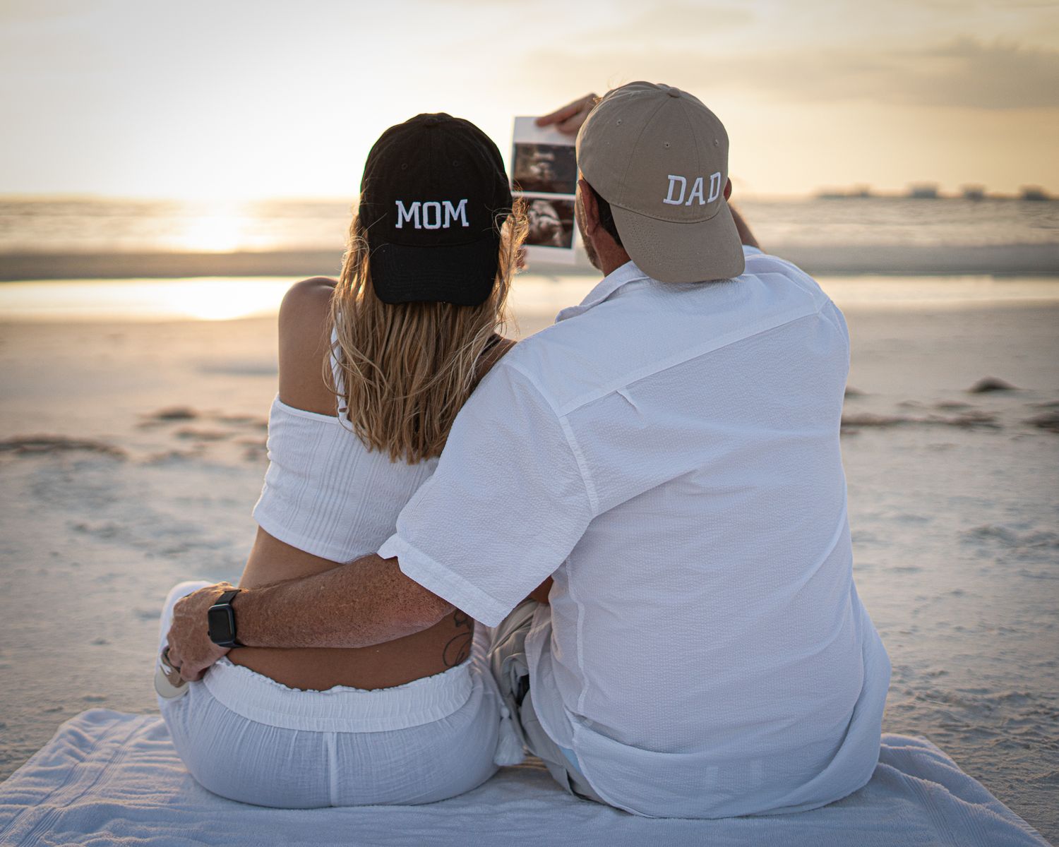 Two people wearing baseball caps sit close together on a beach at sunset.