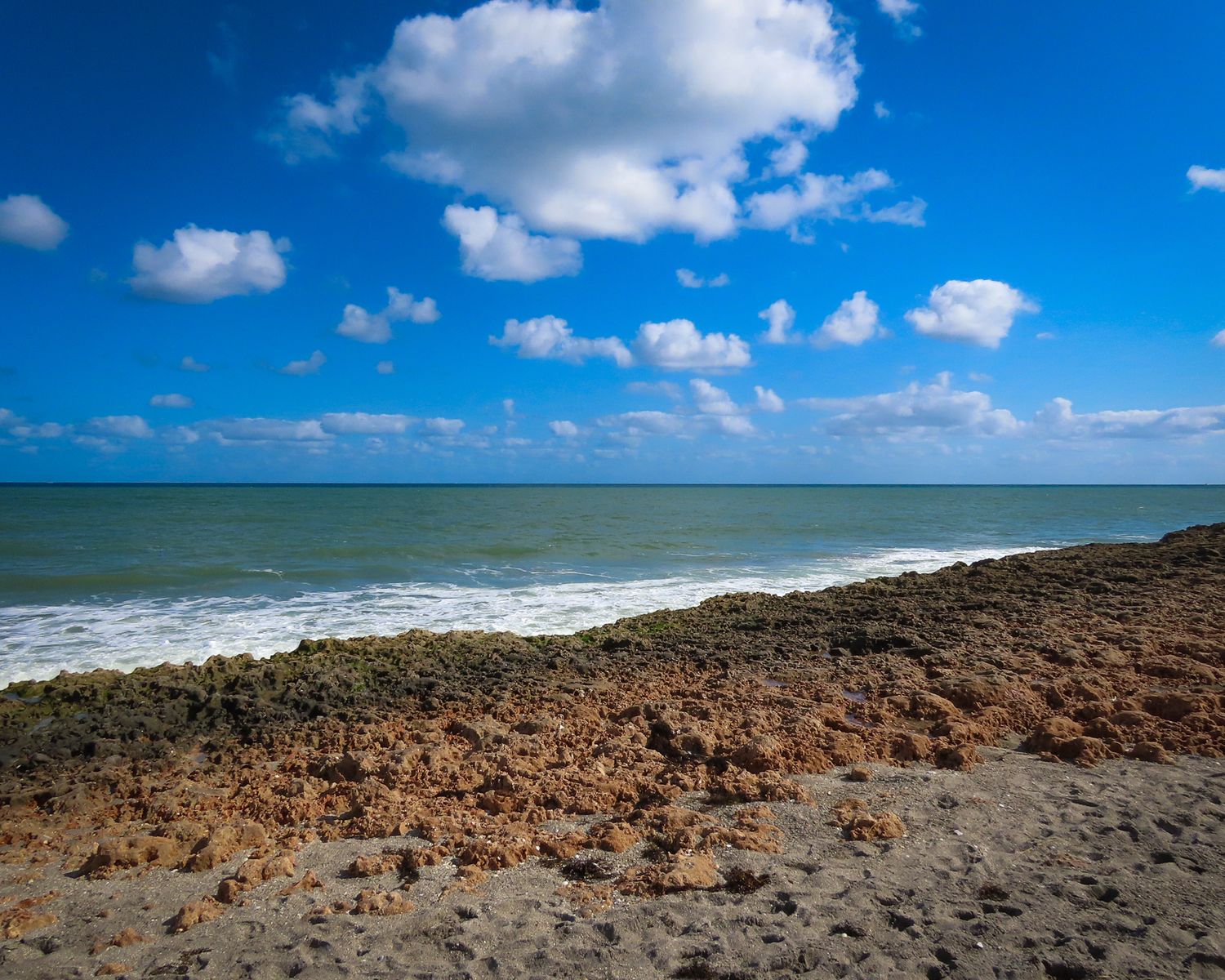 Rocky coastal shoreline under vibrant blue sky with fluffy white clouds and turquoise ocean waters.