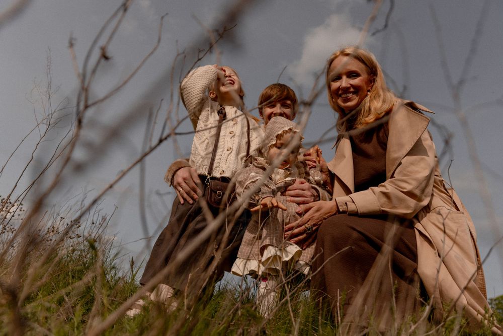 Group of people sitting together in a field wearing coats during autumn while tall grass sways in the breeze.