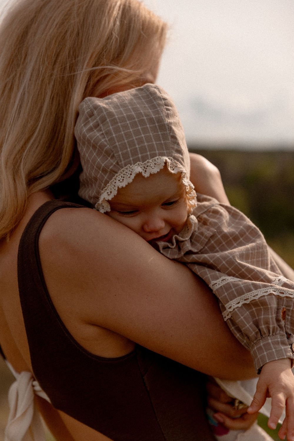 A baby wearing a gray checkered bonnet cuddles on a parent's shoulder in warm sunset lighting.