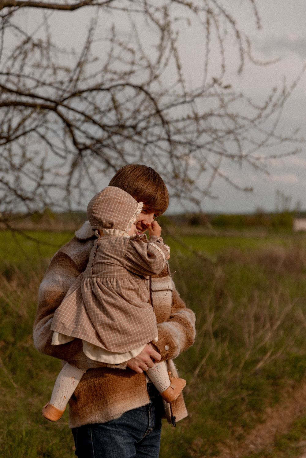 Someone wearing a brown sweater holds a child in a beige dress and bonnet outdoors near bare tree branches.