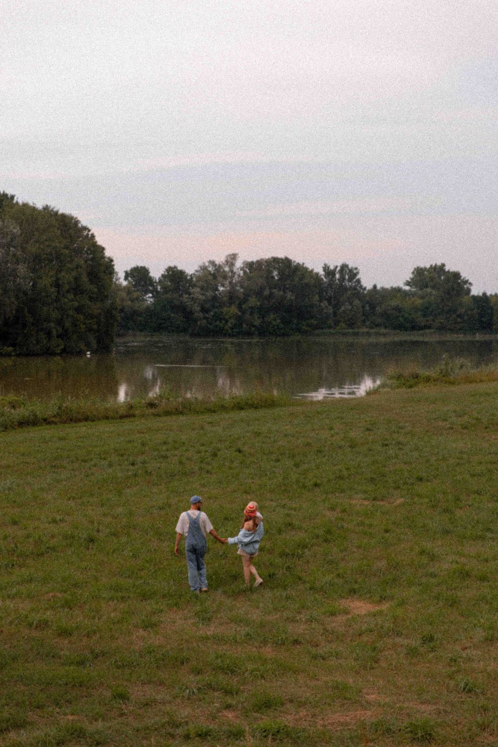 Two people stand on grassy terrain near a tranquil lake surrounded by trees on a misty day.