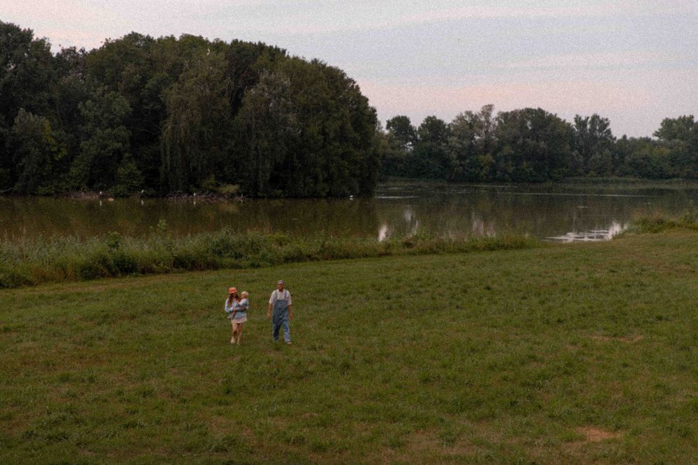 Two people walking through a grassy field near a peaceful lake surrounded by trees at dusk.