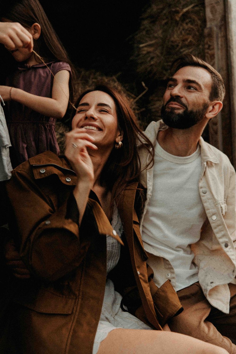 A couple shares a joyful candid moment outdoors while wearing brown jackets in warm sunlight.