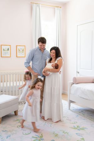 A family plays together in a bright nursery room with floral floor decals and white furniture.