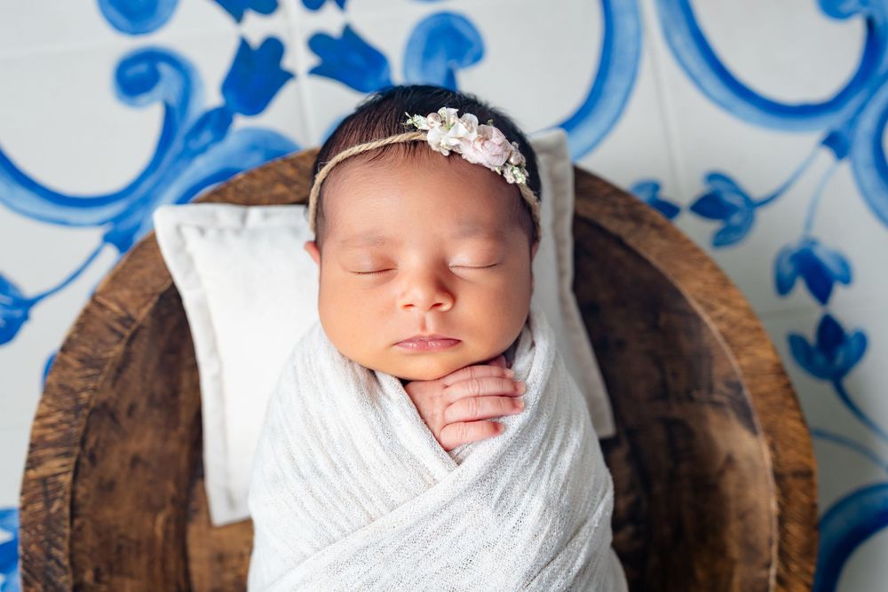 Sleeping newborn swaddled in white wrap with crystal headband resting in wooden bowl against blue floral background.