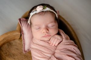 Sleeping newborn swaddled in soft pink fabric resting in wooden bowl with floral headband accessory.