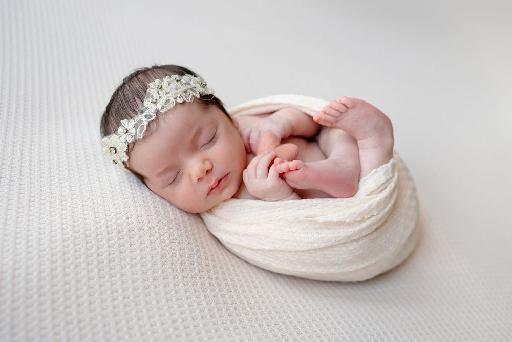 A sleeping newborn wearing a delicate silver headband lies swaddled in soft white fabric on a textured white surface.