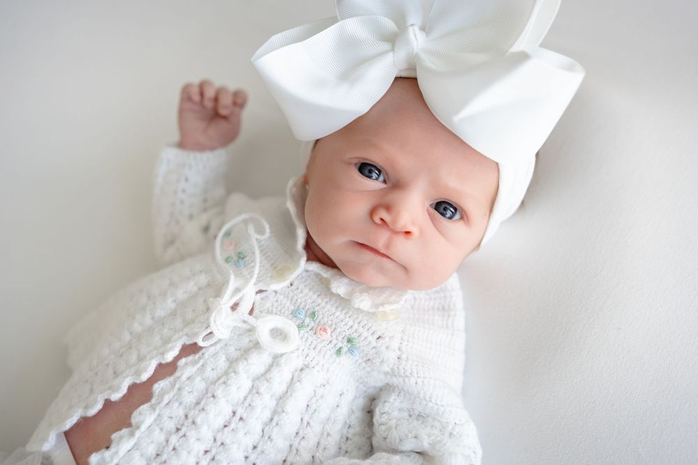 Small baby wearing white knitted sweater and large white bow headband laying against light background.