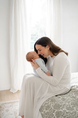 A mother in a white sweater holds and snuggles her newborn baby in a bright, airy room with white curtains.