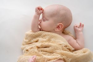 A sleeping baby wrapped in a cozy tan knit blanket stretches peacefully on a light background.