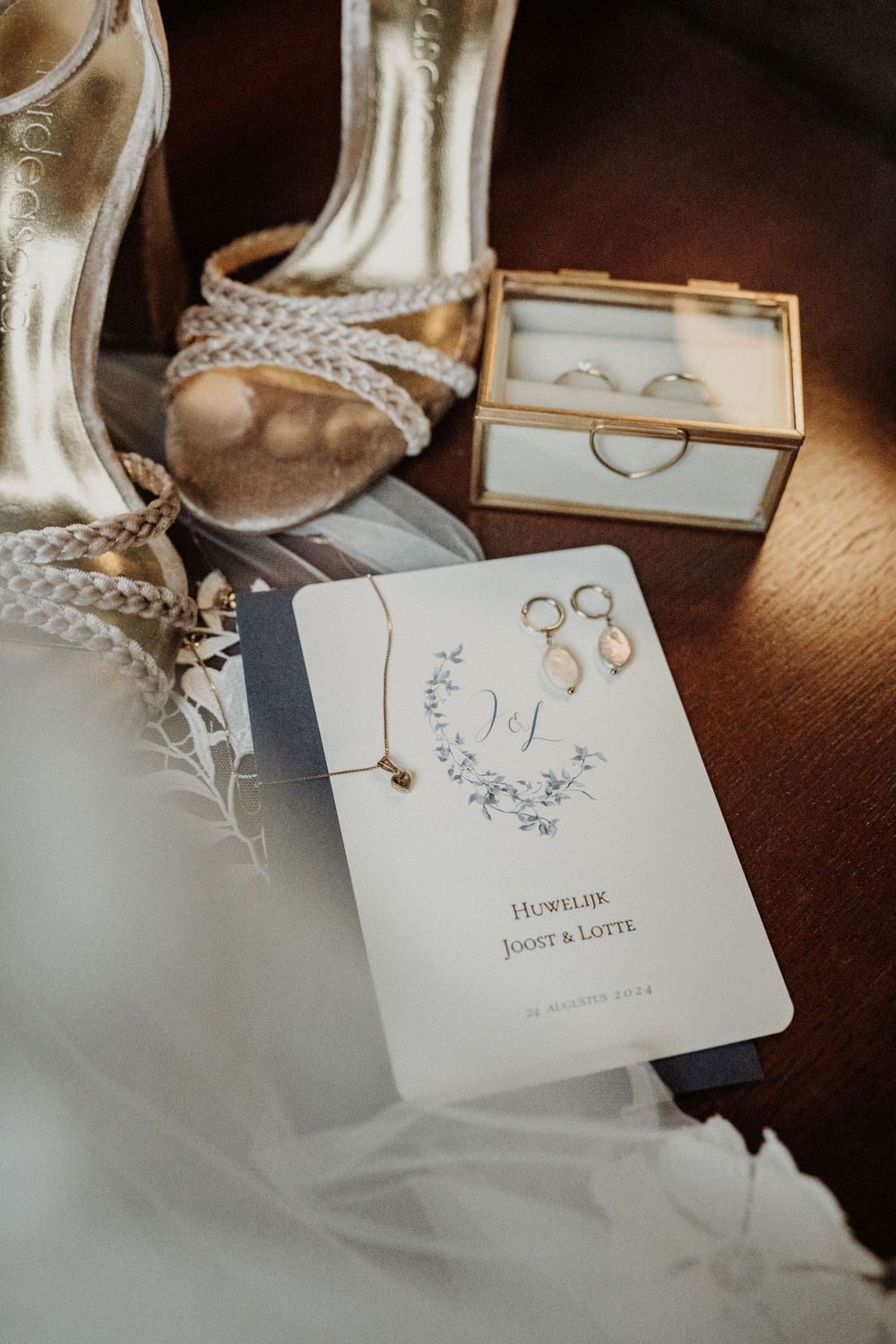 Wedding details including bridal shoes, wedding rings in a box, and an elegant invitation card on a wooden surface.