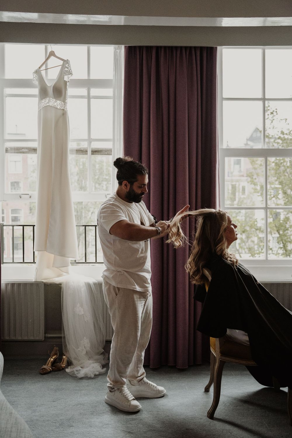 A hairstylist works on a client's hair in a room with a wedding dress hanging by the window.
