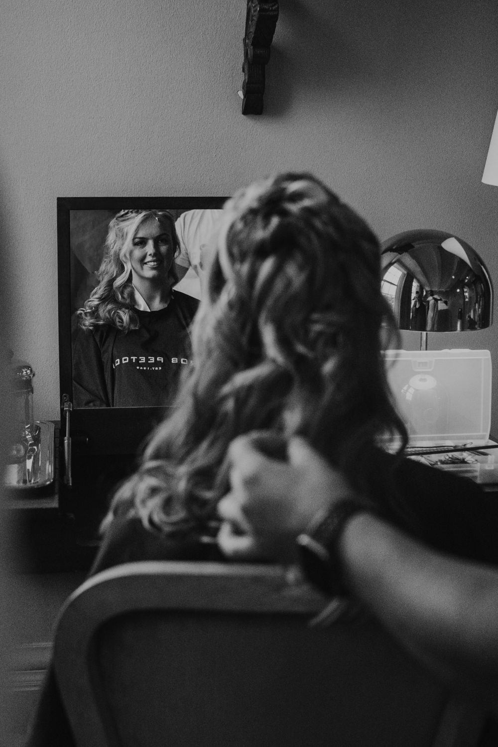 Black and white reflection of a person getting ready at a vanity mirror with a framed photo visible in the background.