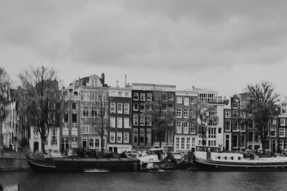 Black and white photograph of traditional canal houses and moored boats along an Amsterdam waterway under cloudy skies.