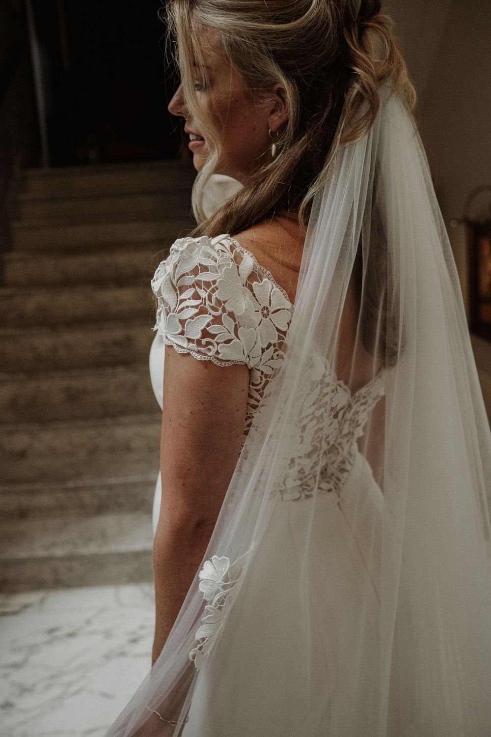 Side view of a bride in a lace cap sleeve wedding dress with flowing veil standing in a rustic setting.