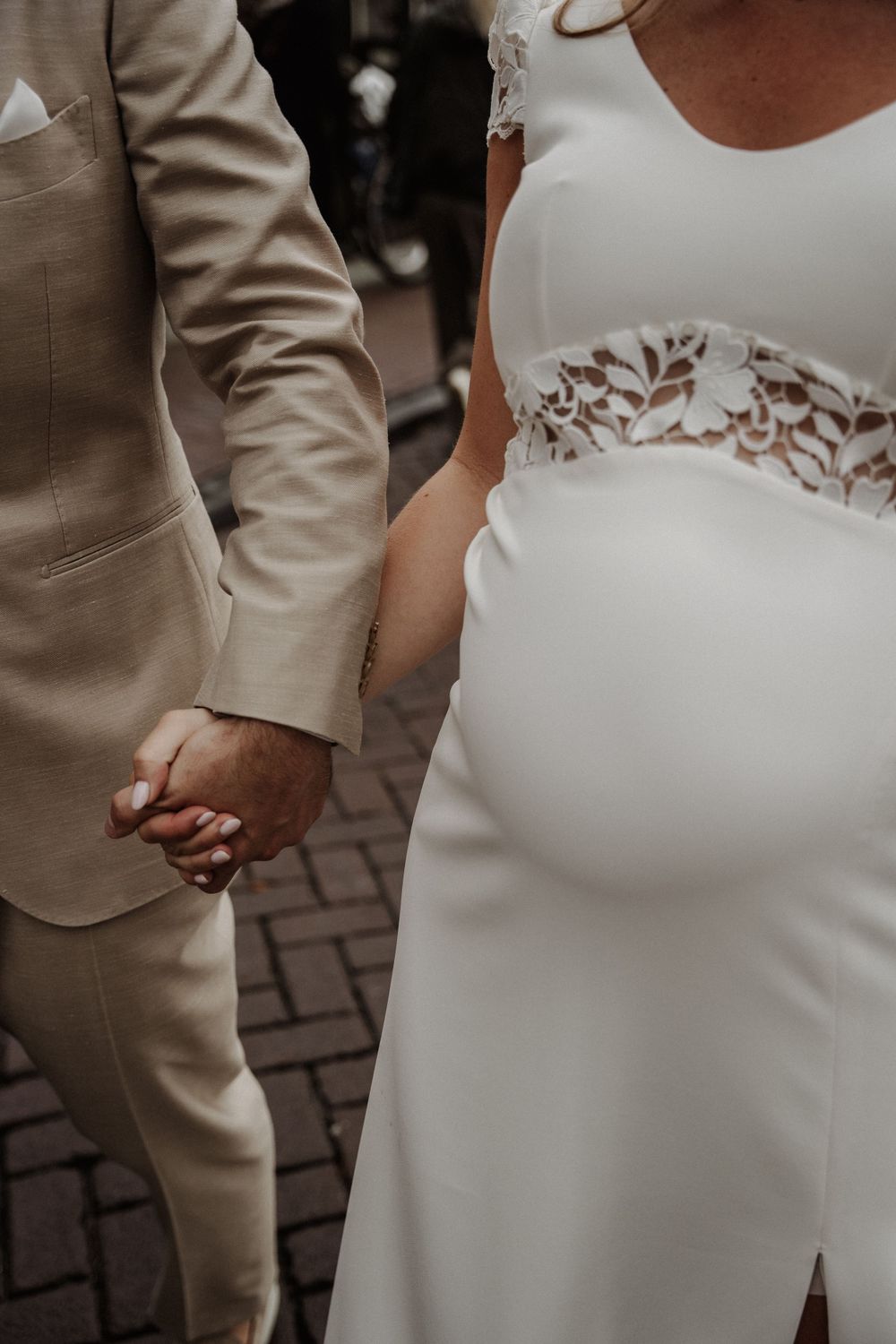 Close-up of couple holding hands at wedding, showing beige suit sleeve and white lace wedding dress details.