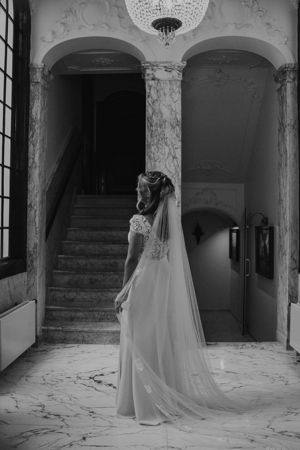 Black and white photo of a bride in an elegant wedding dress and veil standing in a grand marble hallway.