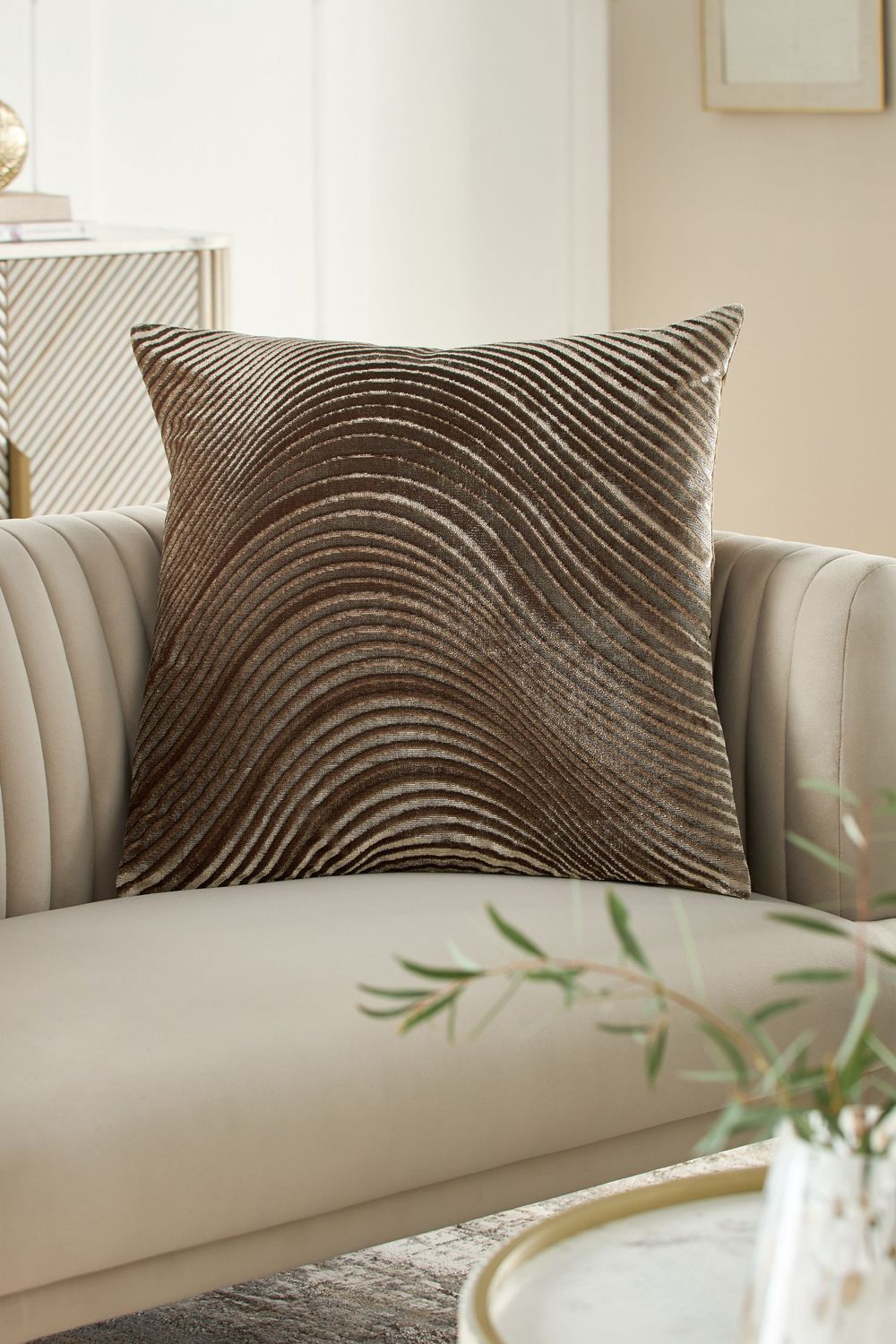 Brown velvet throw pillow with diagonal ribbed texture pattern sits on a beige sofa next to decorative greenery.