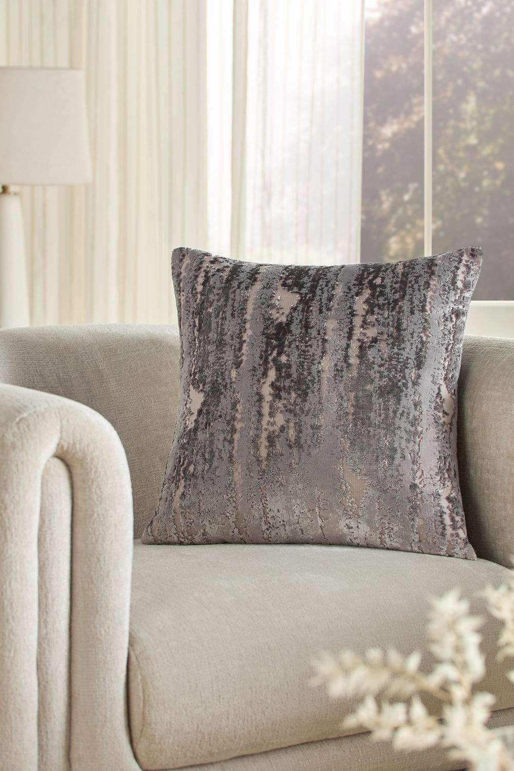 A luxurious gray velvet throw pillow sits on a cream-colored upholstered chair near a sunlit window.