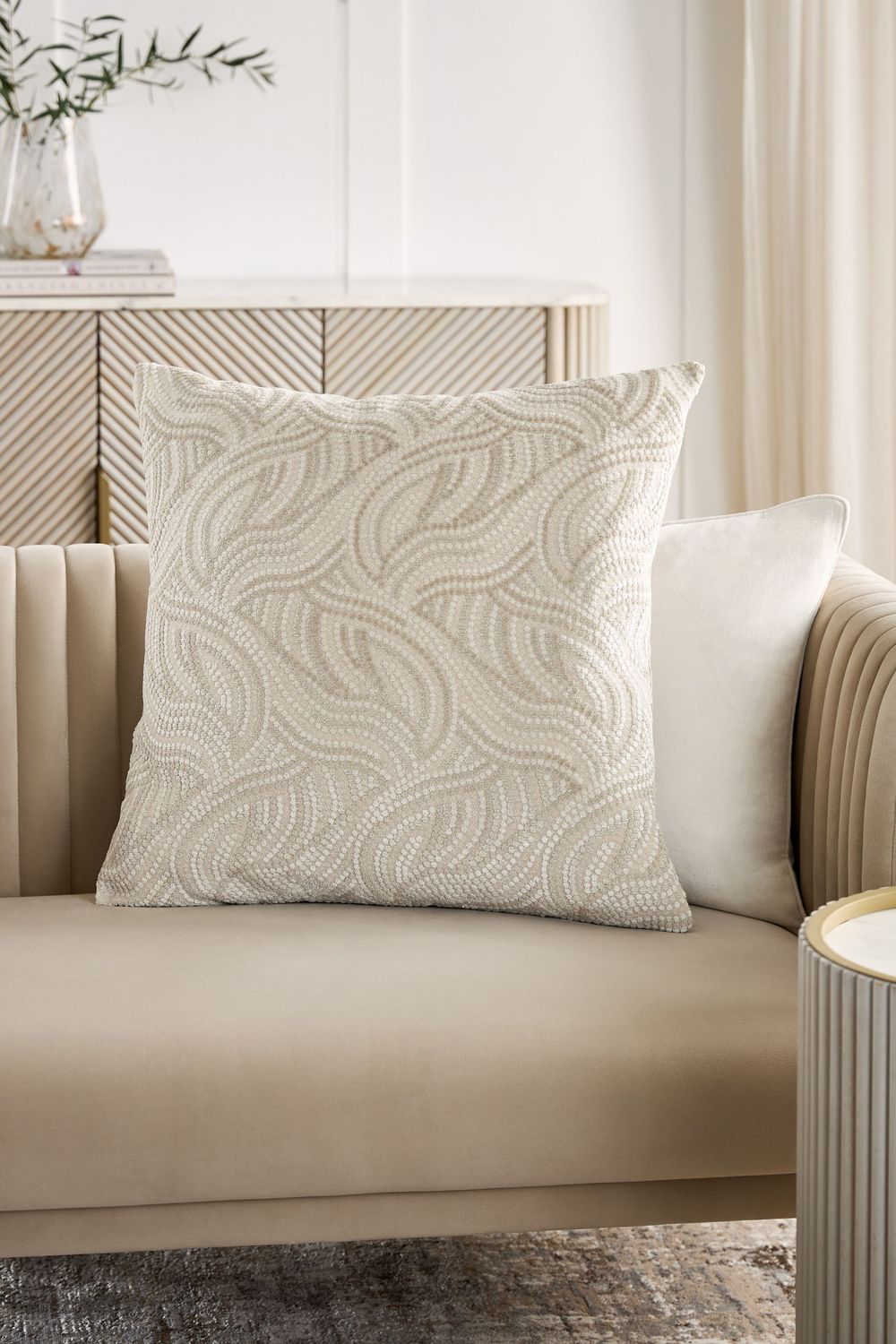 Decorative beige textured throw pillow with swirled pattern on a neutral sofa next to white curtains and accent table.