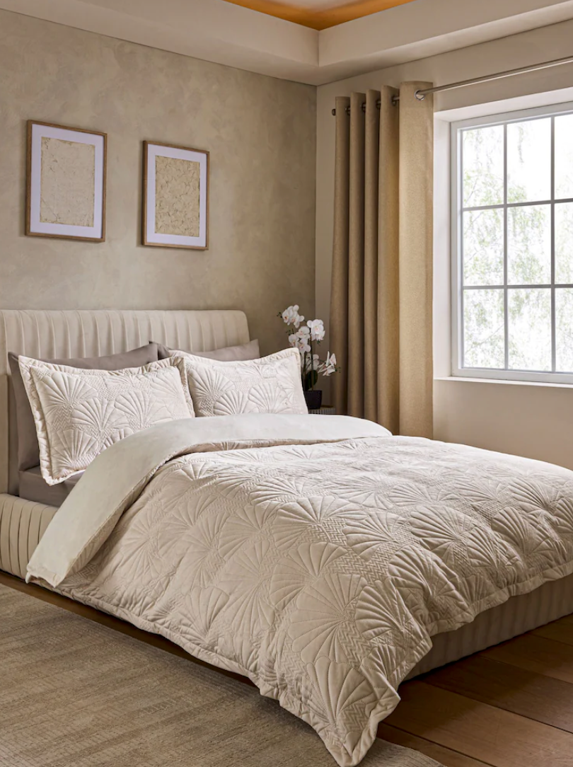 A modern bedroom with beige textured bedding, upholstered headboard, and large window with floor-length curtains.