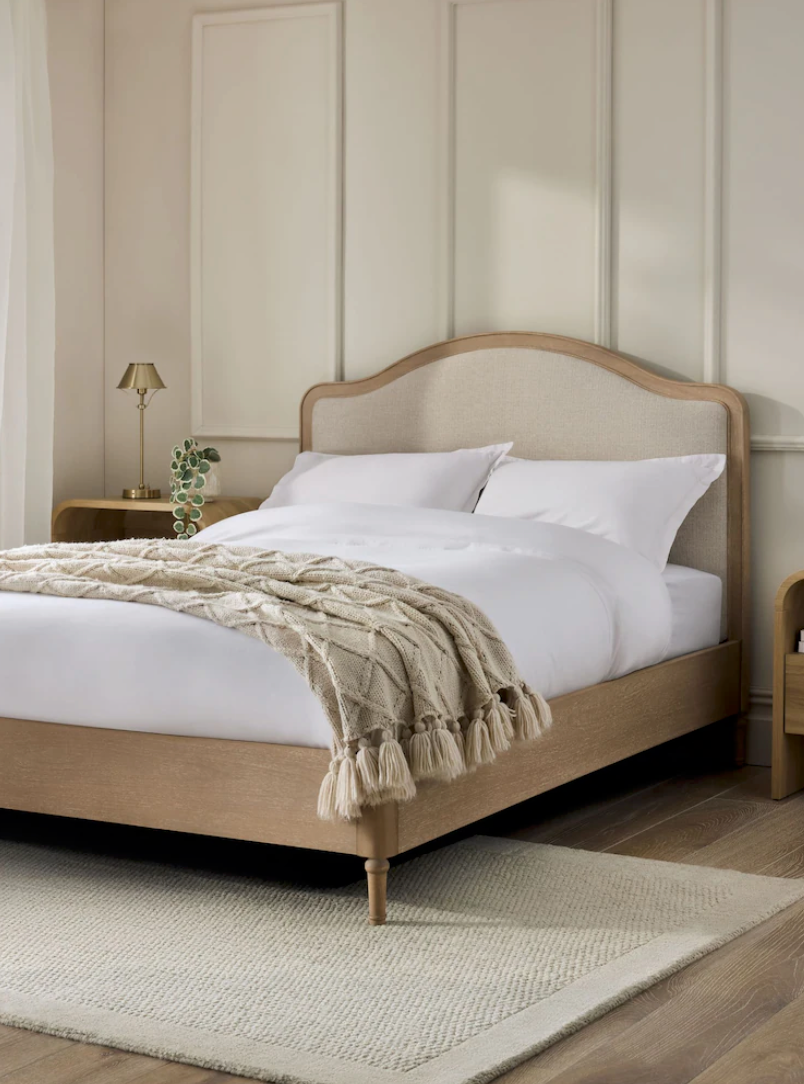 Minimalist beige upholstered platform bed with white bedding and textured throw blanket on light carpet in bright room.