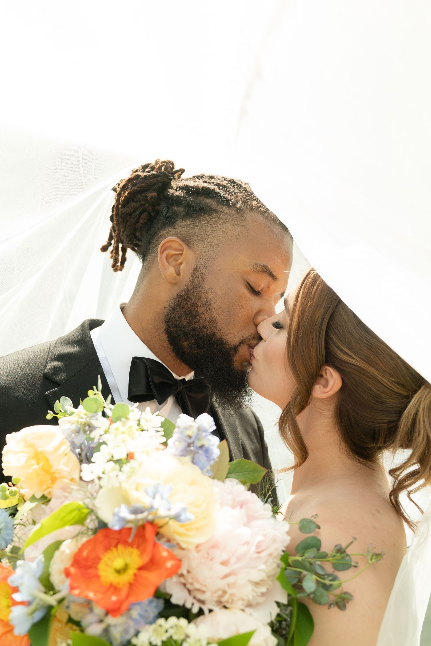 Interracial couple sharing a kiss under a bridal veil, groom in tuxedo, bride holding colorful wedding bouquet.
