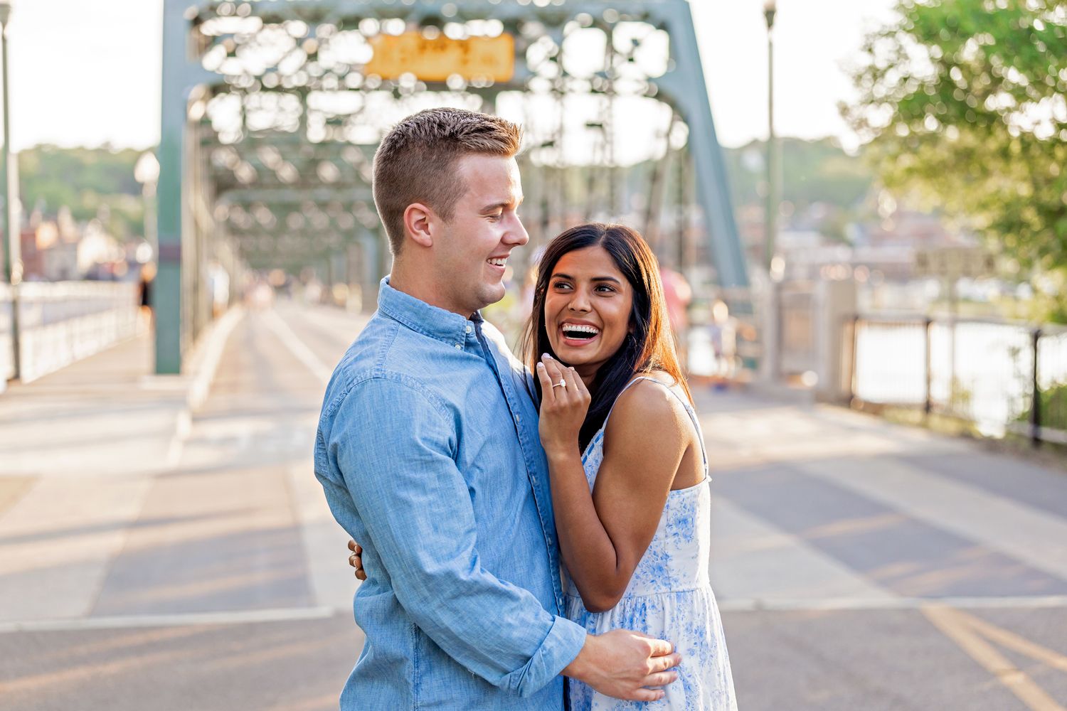 Laughing engaged couple hugging closely on a bridge at sunset after a surprise marriage proposal.