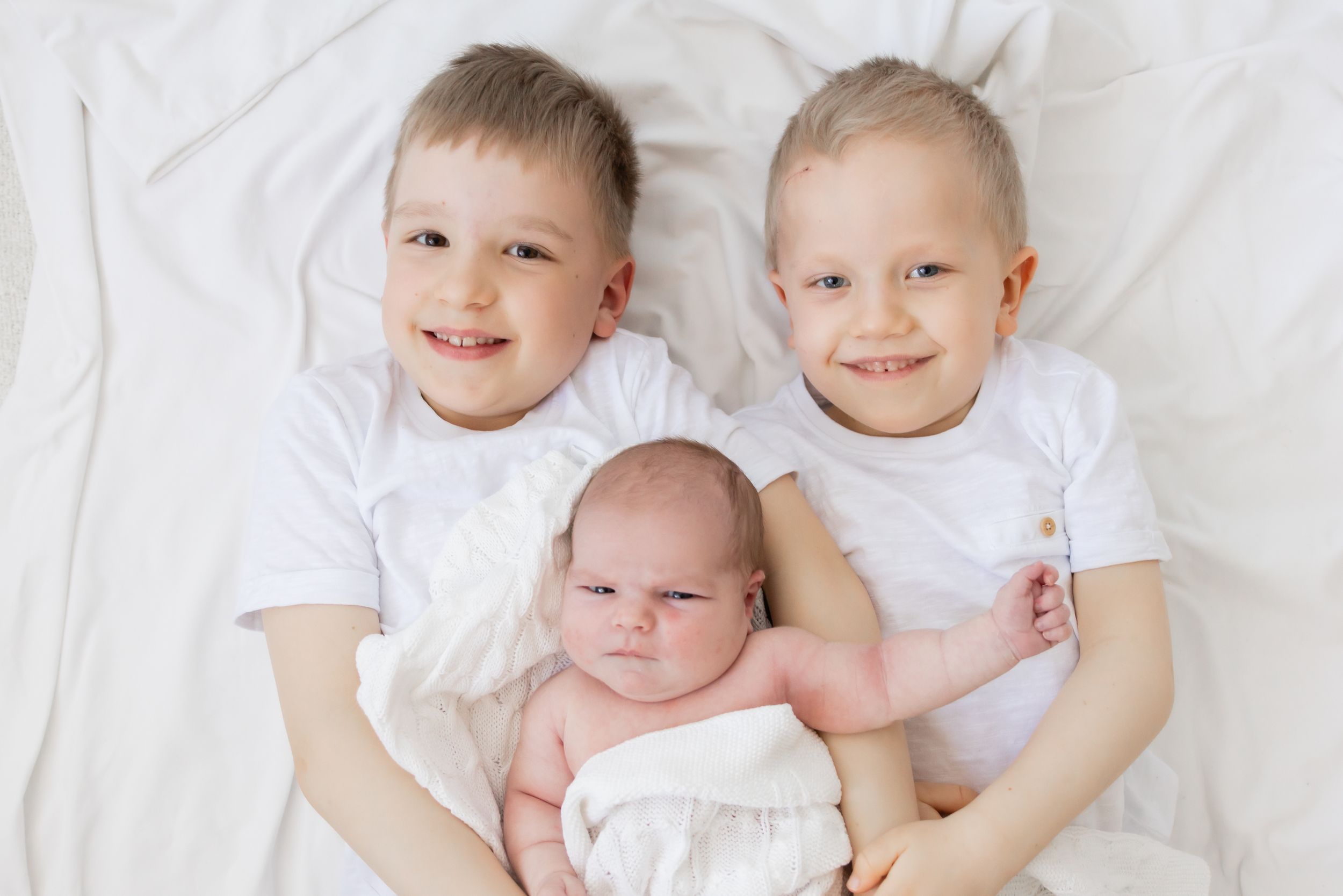 newborn kuvauksessa vanhemmat veljekset pitelee pientä pikkusiskoaan ja hymyilevät